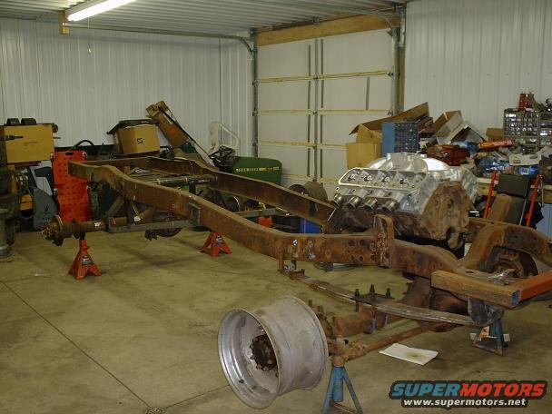 Truck frame on jack stands with a V8 engine block nearby. A large wheel rim is in the foreground