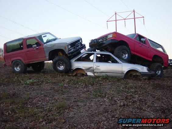 Two lifted Ford Broncos are stacked on a silver sedan that is crushed on the ground