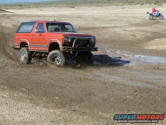 Orange Ford Bronco with aftermarket bumper driving through mud; ATV in background