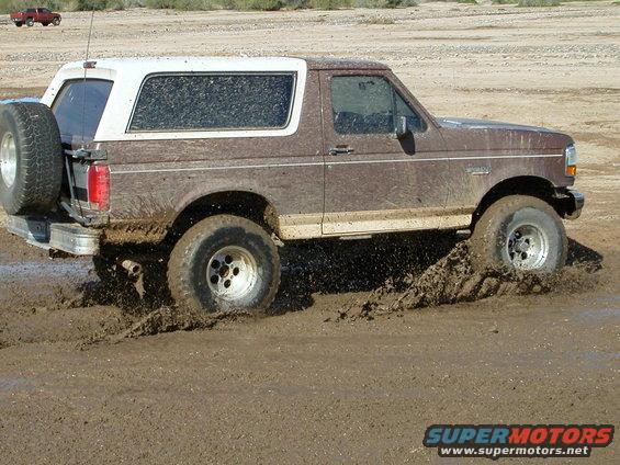 Brown and white Ford Bronco stuck in mud, kicking up dirt. Another red truck is visible in the distance