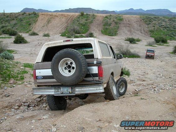 Ford Bronco with spare tire on back, driving on dirt road with another vehicle in distance