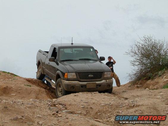 Dark gray Ford Ranger pickup truck navigating a dirt trail with two people visible inside and one standing nearby