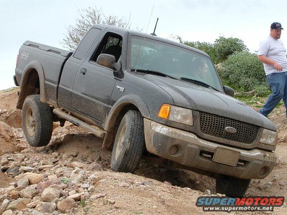 Black Ford Ranger pickup truck driving over rocks on a dirt trail. A man walks behind it
