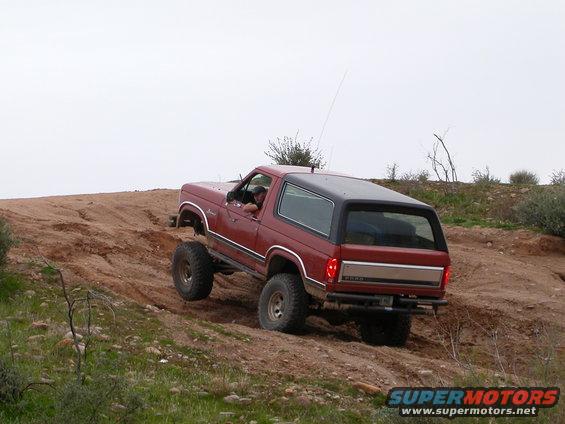 Red Ford Bronco with lifted suspension climbing a steep dirt hill, rear 3/4 view