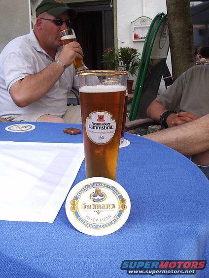 Man in green hat drinks beer next to a glass of Neumarkter Lammsbrau and a Gutmann coaster on a blue tablecloth