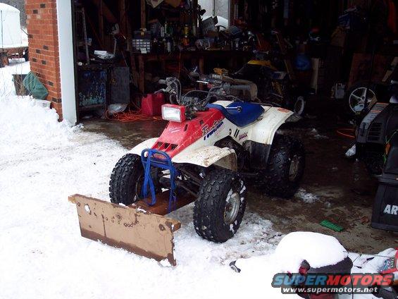 Red and white ATV with a front plow attachment, parked in a snowy garage. Blue straps secure the plow