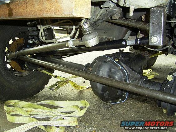 Close-up of vehicle's front axle, steering linkage, and suspension components with yellow tie-down straps on the ground