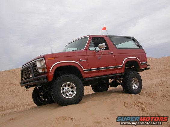 Red Ford Bronco with black trim and a black roof, lifted on large tires, driving on sand dunes