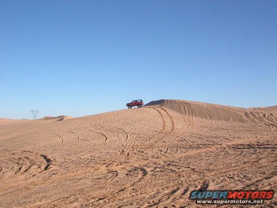 Red 1984 Ford Bronco driving on a sand dune with tire tracks