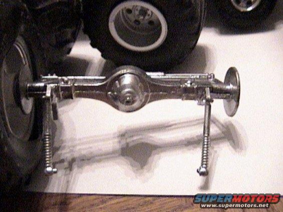 Model car rear axle assembly with springs and shocks, chrome finish, next to a tire
