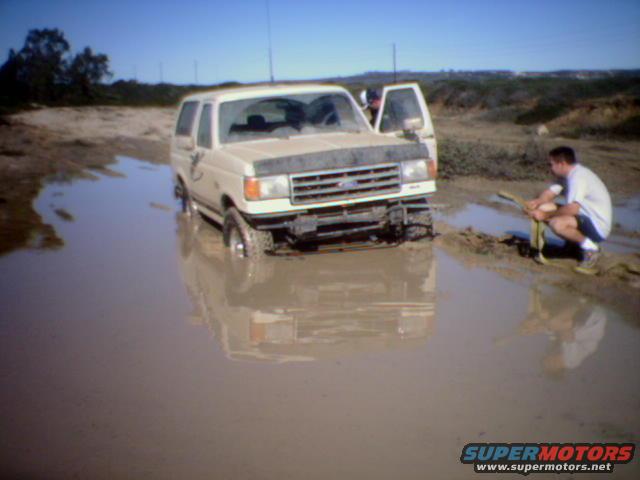 1990 Ford Bronco stuck in a mud puddle with a person nearby holding a tow strap