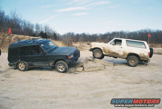 White Ford Bronco climbing concrete blocks, black SUV beside it