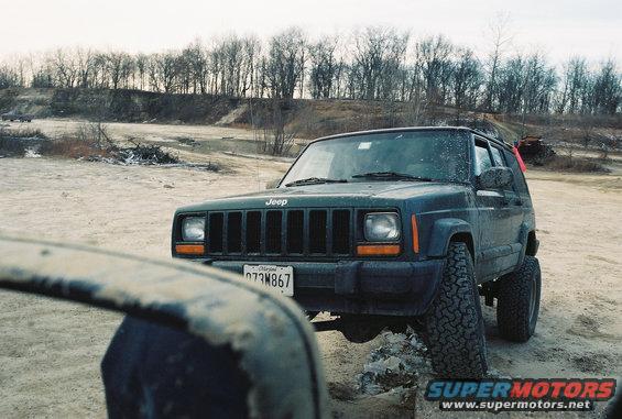 Dark green Jeep Cherokee XJ parked on dirt, front 3/4 view, with a dirty mirror in the foreground