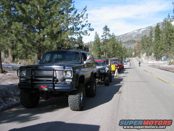 Dark blue SUV with brush guard and roof rack, front view. Several other off-road vehicles in line