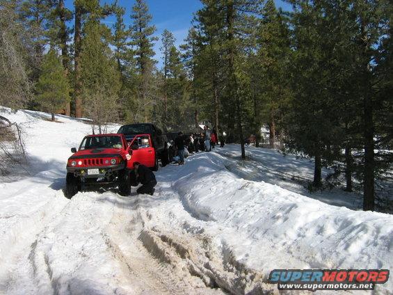 Red off-road vehicle with door open, people around it in snow. Another black vehicle behind it