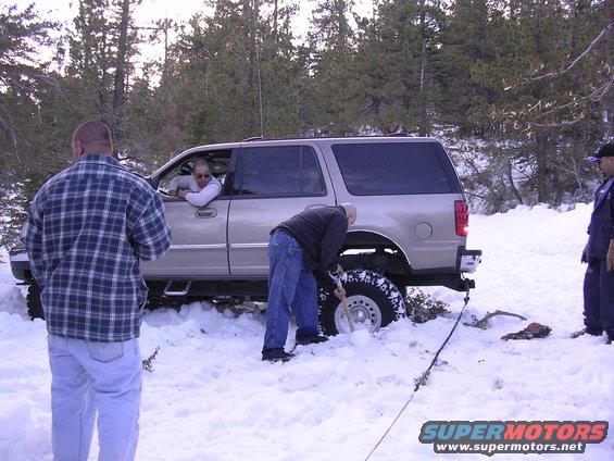 SUV stuck in snow, people digging around the front wheel. Driver in window