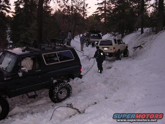Several off-road vehicles partially buried in snow on a wooded trail, with people attempting to pull one out with a rope