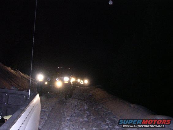 Several trucks driving on a dirt road at night, headlights illuminating the path