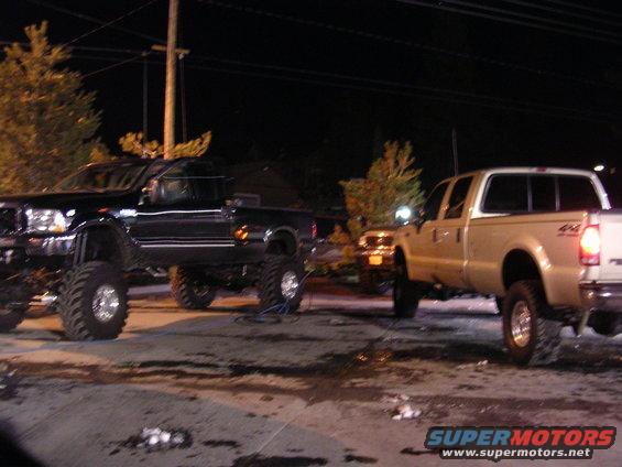 Black pickup truck on left, gold pickup truck on right, both lifted with large tires. Night scene