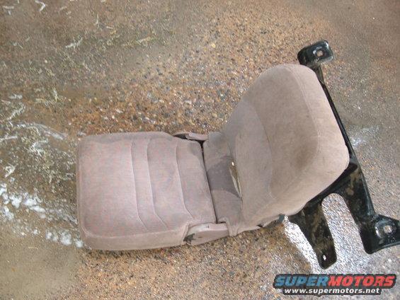 Tan fabric car seat with black metal mounting brackets, on a wet, gravelly surface