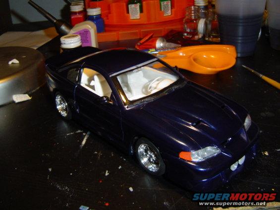 Dark purple model car with white interior on a cluttered workbench. Supermotors.net logo visible