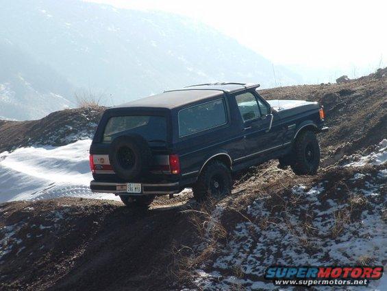Dark blue Ford Bronco climbing a dirt hill with snow patches. License plate reads 246 MTD