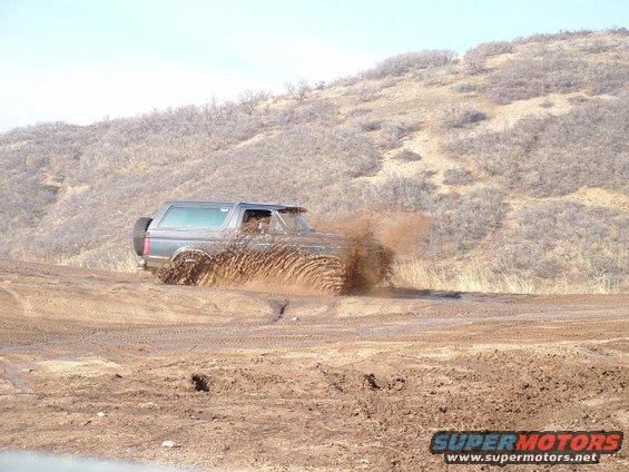 Gray Ford Bronco driving through mud, kicking up a large spray. Spare tire visible on rear
