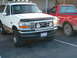 1996 Ford Bronco - A white Ford Bronco is parked next to a red pickup truck, showing its front grille and lights.