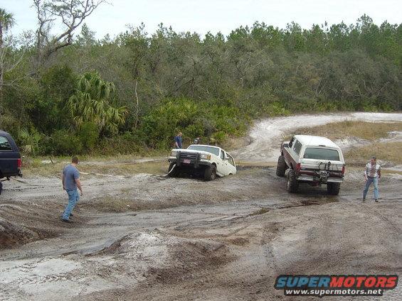 White Ford Bronco stuck in mud, red and white Ford Bronco driving past, men standing nearby