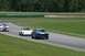 1965 Ford Mustang - Three race cars, including a 1965 Ford Mustang, compete on a paved track with grassy verges.