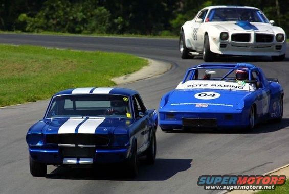 Blue 1965 Ford Mustang with white racing stripes on a track, viewed from the front