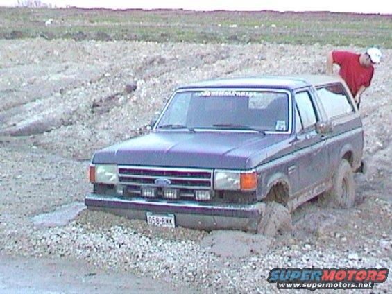 Dark blue Ford Bronco stuck in mud, front driver's side wheel submerged, person in red shirt leaning over roof