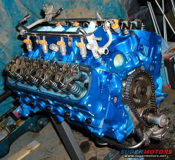 Blue engine block with fuel injectors, rocker arms, and timing chain visible. Engine is on a stand