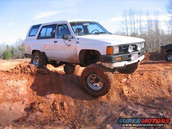 White Toyota 4Runner with off-road tires climbing a dirt hill. Another vehicle is visible in the background