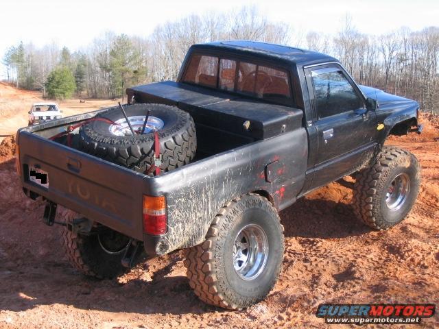 Black Toyota pickup truck with off-road tires, spare tire in bed, and a toolbox
