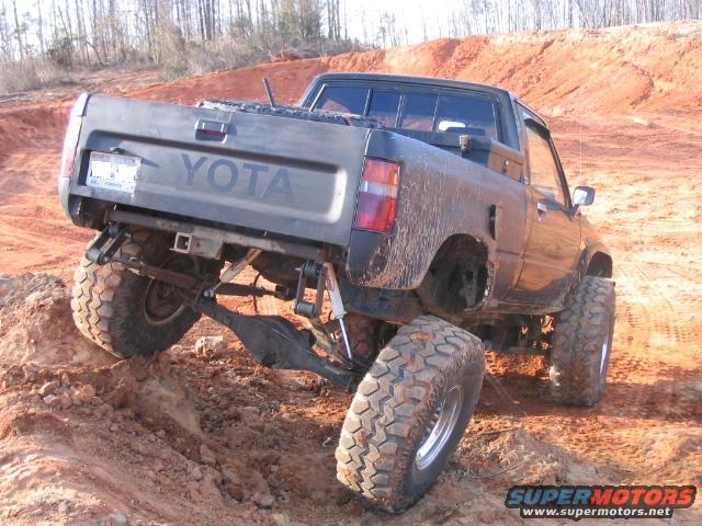Dark gray Toyota pickup truck with large off-road tires climbing a dirt hill. Body style suggests 1980s