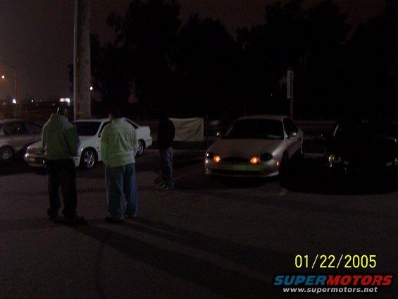 Silver sedan with orange turn signals parked at night. Two people stand nearby. Another car is visible to the right