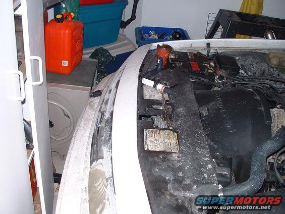 Car engine bay with battery, fan, and hoses visible. Orange gas can and blue bin in background