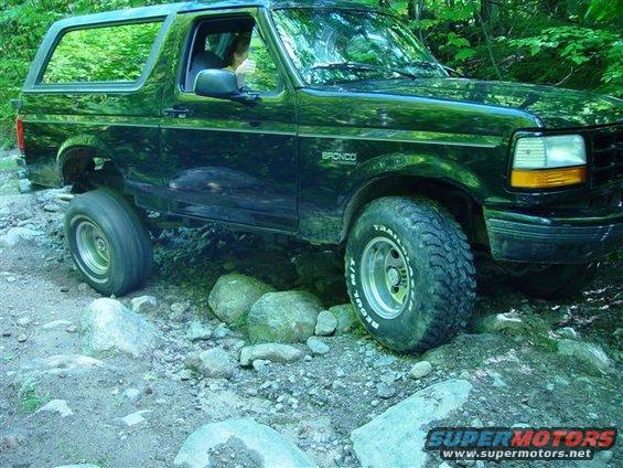 Black Ford Bronco driving over rocks on a wooded trail. Bronco badge visible on fender