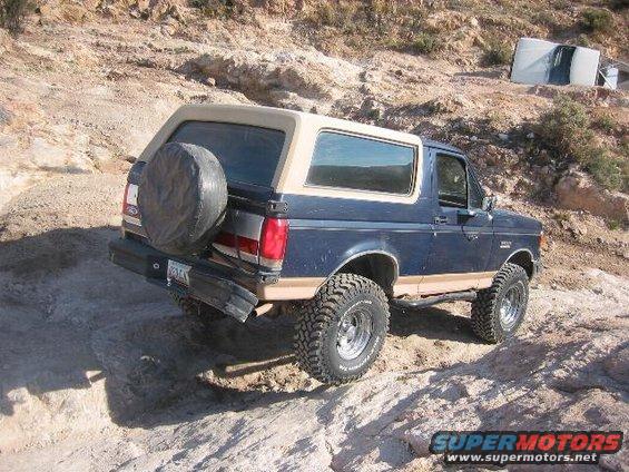Blue 1989 Ford Bronco with tan top driving on a rocky trail, spare tire on back