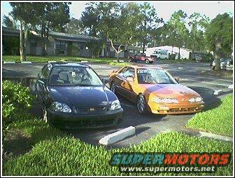 Black car and orange car parked in a lot. The orange car has racing stripes