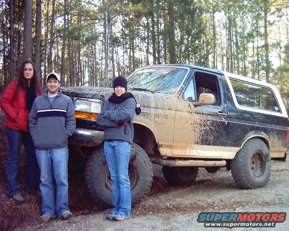 Mud-covered black and white Ford Bronco, 3/4 front view, with three people standing beside it