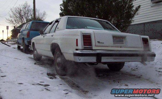White Ford Crown Victoria on snowy road, blue pickup truck behind it