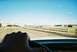 2000 Ford Crown Victoria - View from inside a vehicle as it drives on a dirt road towards a building and sports fields under a clear blue sky.