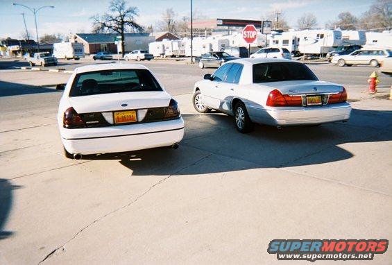 Two white Ford Crown Victorias parked on pavement, one slightly ahead of the other. Rear view of both