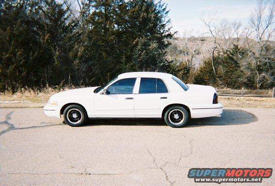 Side view of a white 2000 Ford Crown Victoria parked on a road with trees in the background