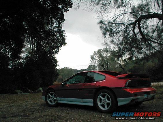 Red 1986 Pontiac Fiero parked on dirt road with trees and overcast sky