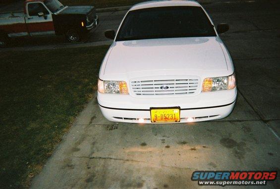 Front view of a white 2000 Ford Crown Victoria parked on a driveway, with a red and black pickup truck in the background