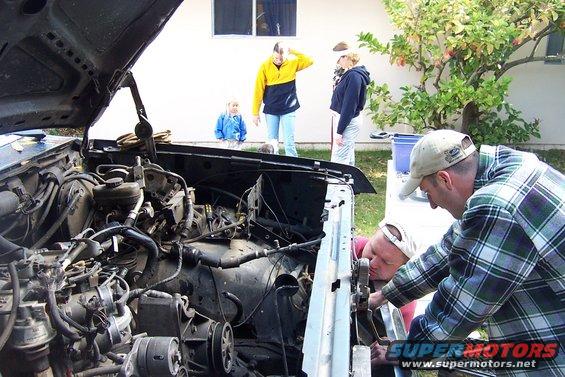 Two men working on an open car engine bay; two children and an adult watch in the background