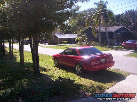 Red 1991 Mazda MX-6 coupe parked on a grassy lawn, rear 3/4 view. Sun glinting off the spoiler
